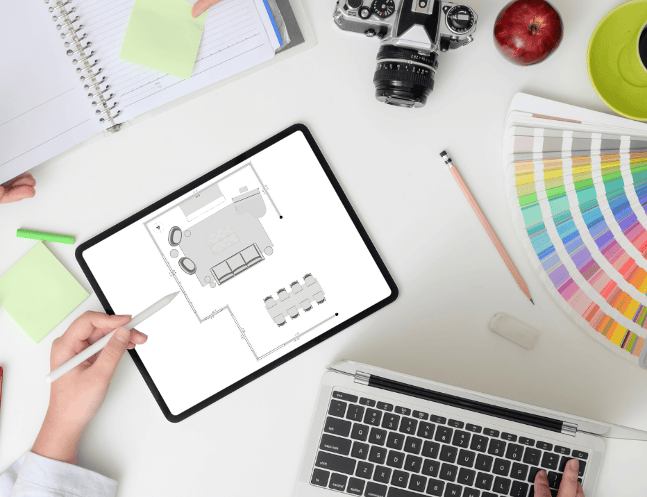 An overhead view of a workspace with a person using a stylus on a tablet showing floor plans, surrounded by a laptop, camera, color swatches, and stationery.
