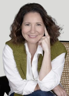 This image features Catherine Severson, founder of design-chat, an e-design interior design firm, smiling gently at the camera while resting her chin on her hand, wearing a white shirt and olive sweater wround her shoulders.