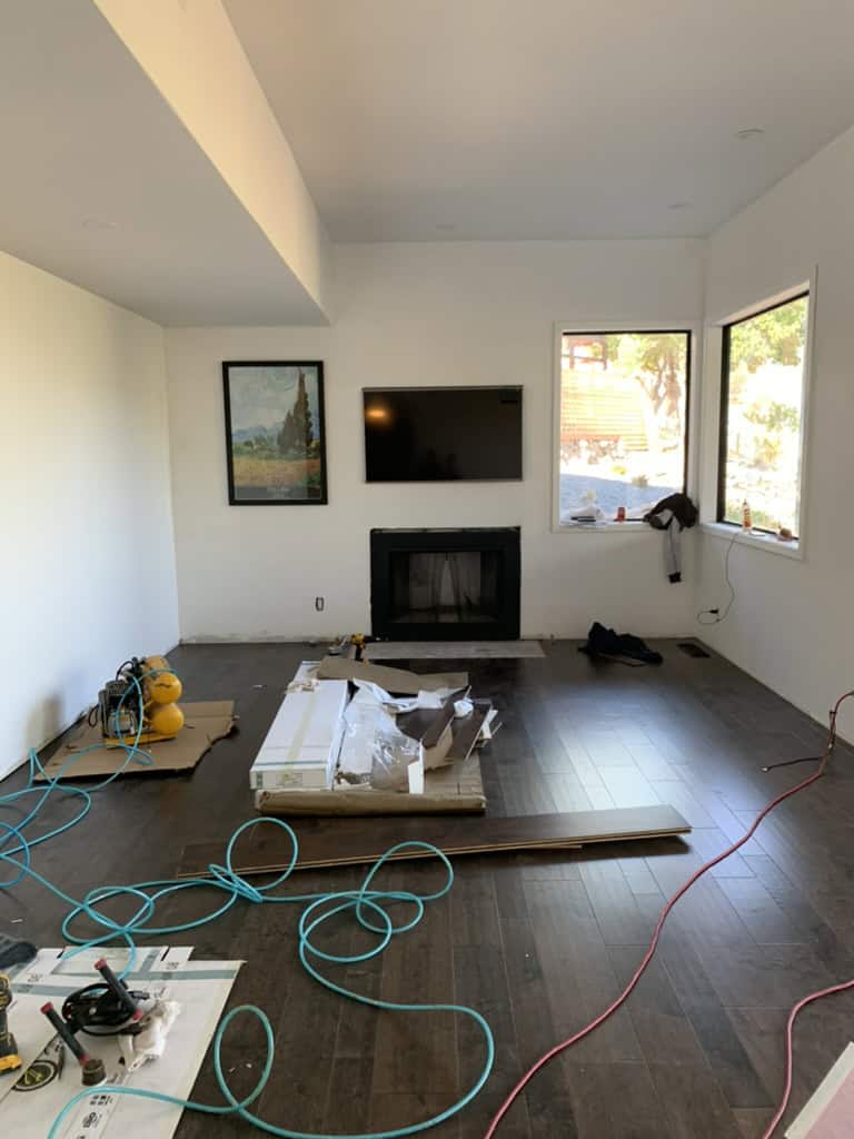 A room under renovation with new flooring being installed. Tools, cords, and unpacked materials are scattered around. Sunlight enters through the windows.