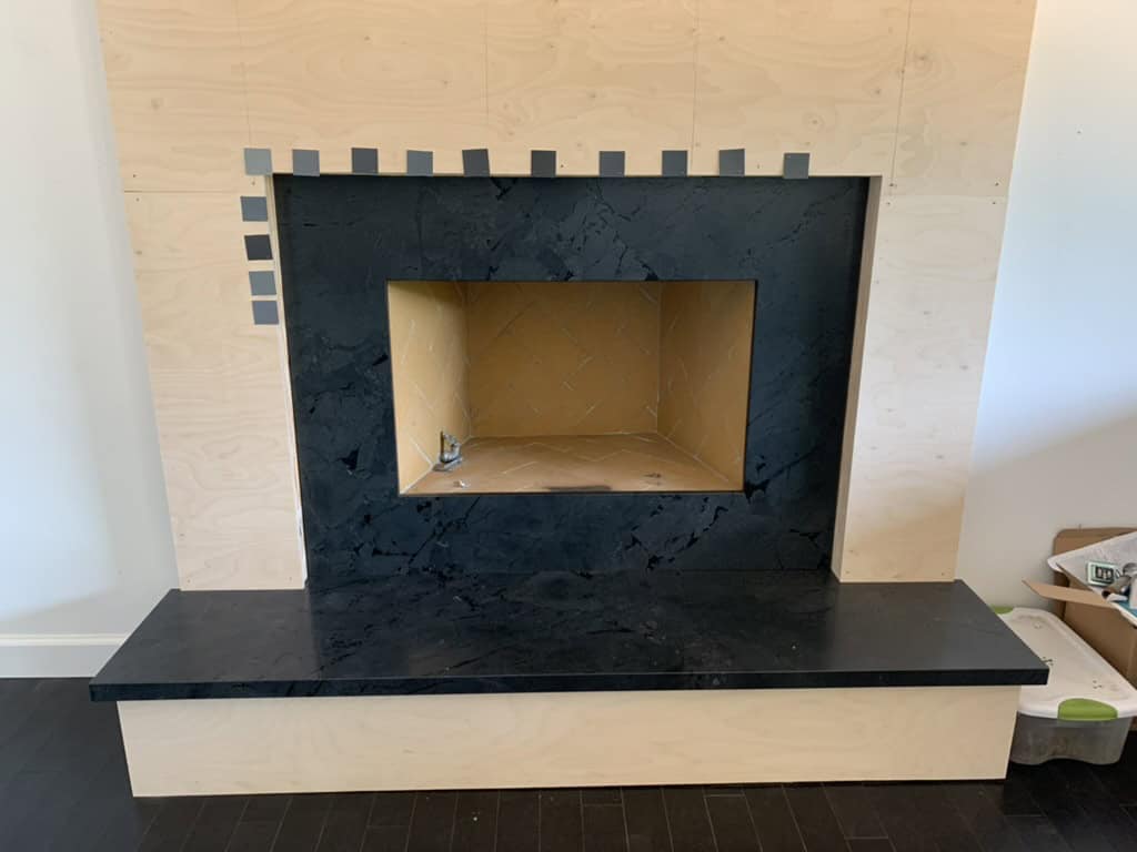 A modern fireplace with black marble surround and hearth, set against a plywood wall, is under construction with multiple paint chips surrounding it as part of a benjamin moore paint color palette package with design-chat.