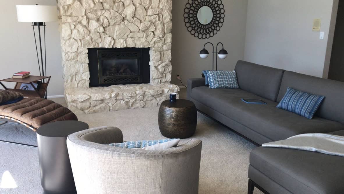 A modern living room with a stone fireplace, a grey sectional sofa with blue pillows, chairs, a lamp, and a round mirror on the wall - the completed virtual room makeover by design-chat.