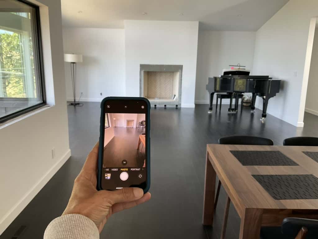 A person is taking a photo during their text-chat and virtual room makeover with design-chat. The image shows a spacious room with a dining table and a black grand piano, using a smartphone, with natural light coming through windows