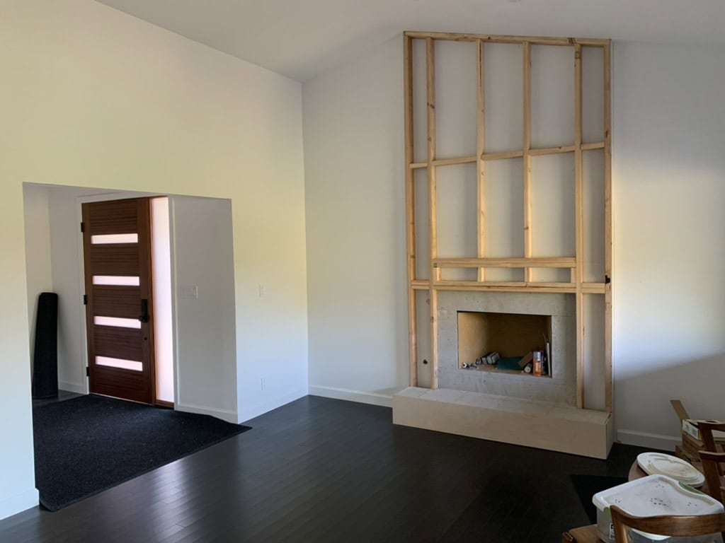 The image shows an empty room with white walls, a dark floor, a wooden door, and a fireplace with an unfinished wooden frame around it. This room is a blank slate for a benjamin moore paint color palette from design-chat.