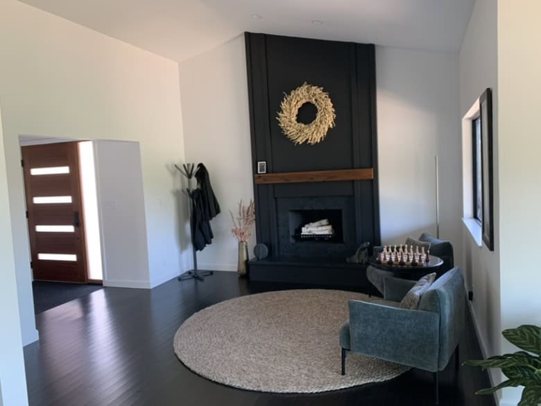 A modern living room with a black fireplace, two teal chairs, a round rug, and a decorative wreath above the mantel. Natural light streams in.