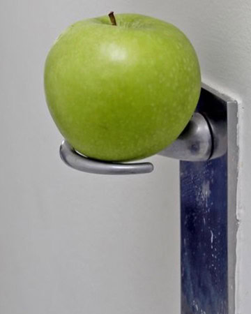 A green apple is perched on a metallic door handle attached to a white wall, creating a quirky and unconventional visual juxtaposition.
