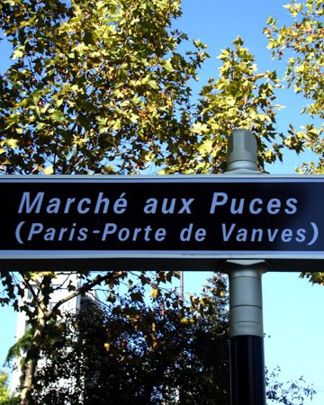 The image shows a signpost that reads "Marché aux Puces (Paris-Porte de Vanves)" against a background of green tree leaves and a clear blue sky.