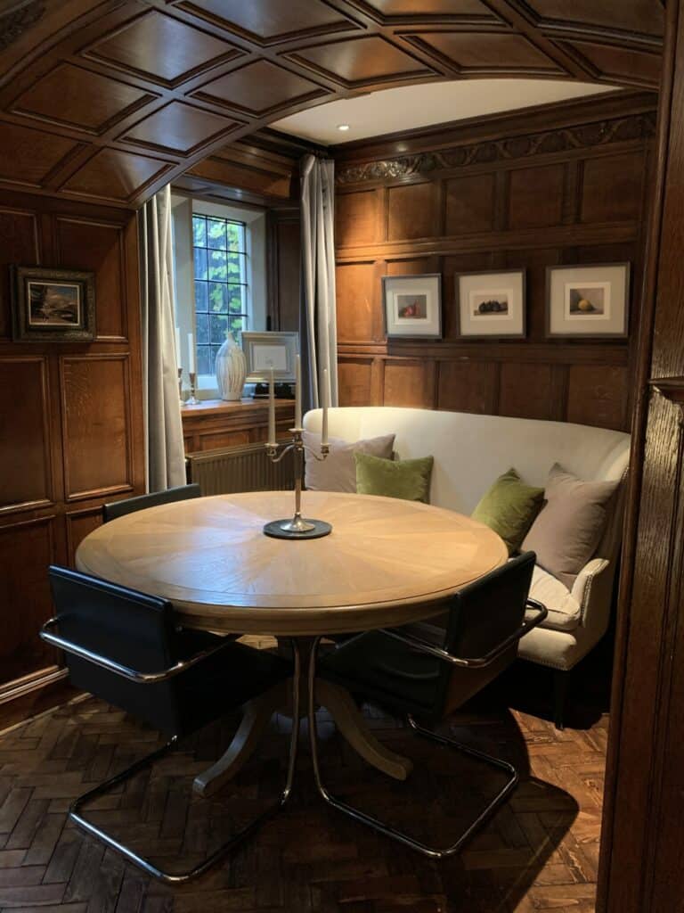 The image shows a cozy, wood-paneled room with a circular table, modern chairs, a comfortable bench with pillows, artwork on the walls, and a window. A completed room design with virtual interior designer at design-chat.