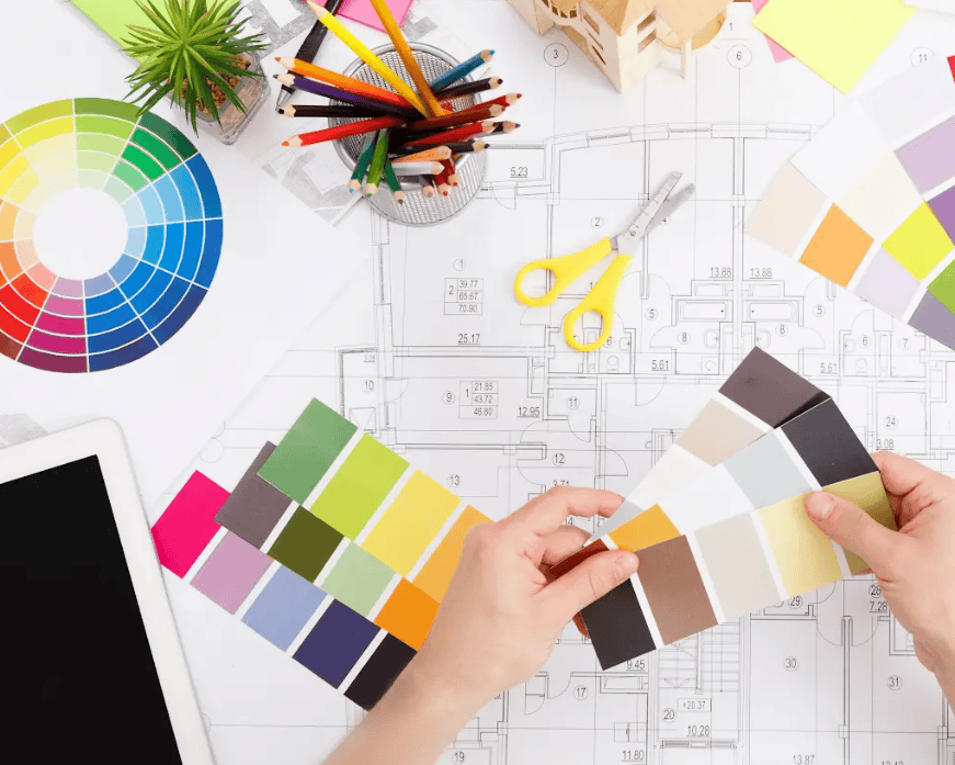 An overhead view of a workspace with architectural plans, color samples, drawing tools, a laptop, and a person's hands comparing color swatches. A picture of working with a virtual interior designer.