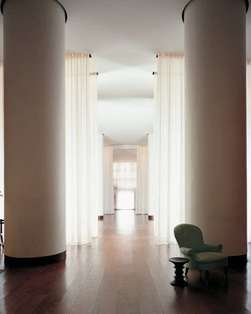 A minimalist corridor featuring tall cylindrical columns, white sheer curtains, dark wood flooring, and a solitary mint green armchair to the left foreground.