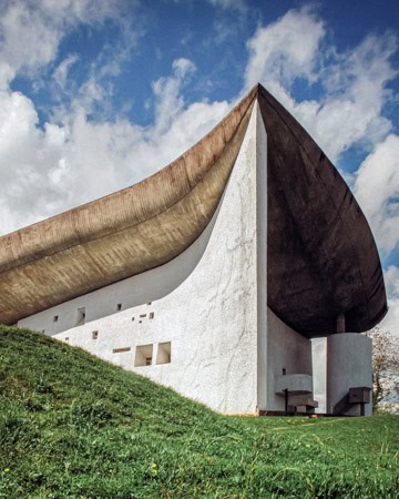 An architectural structure with a unique wavelike roof, set against a cloudy sky, sits atop a grassy hill. Its modern design exhibits organic forms.
