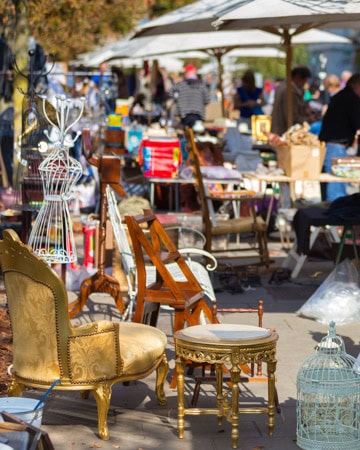 An outdoor flea market scene with various items displayed, including chairs, tables, and decorative objects. People browse in the background under a clear sky. Part of a collection of inspirational images from Catherine Severson, the virtual interior designer who created design-chat.