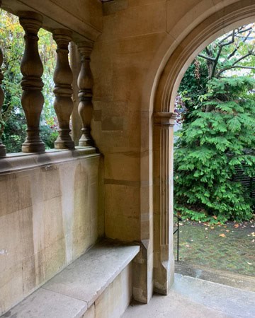 An archway with ornate stone balusters leads to a garden with greenery. A stone bench is integrated into the wall beside the archway.