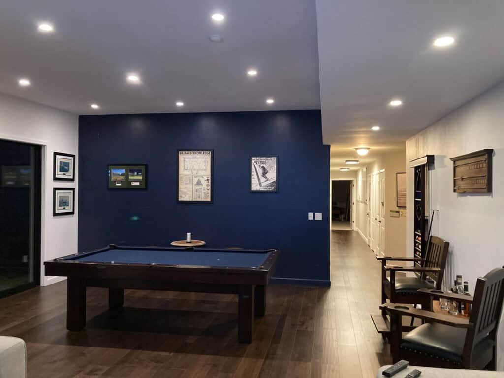 The image depicts a modern game room with a pool table, framed artwork, wood flooring, recessed lighting, chairs, and decorative items on the wall.