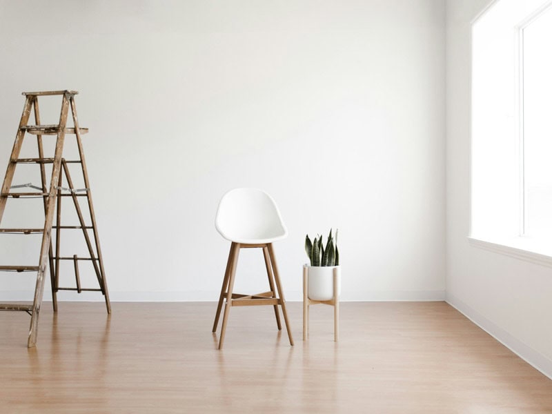 A minimalist room with a wooden floor features a white chair, a worn wooden ladder, and a small white planter with green plants.