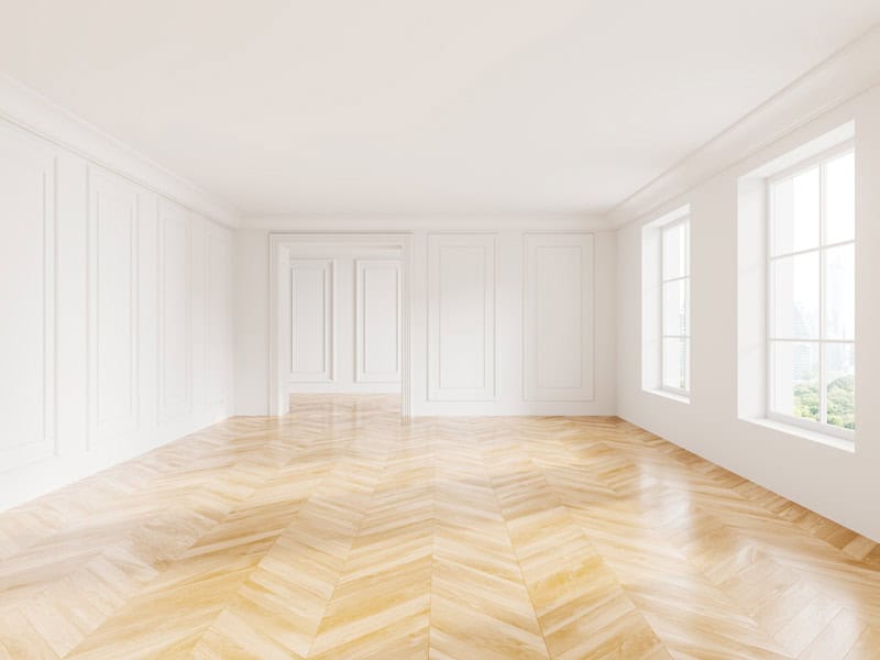 The image shows a bright, empty room with herringbone parquet flooring, white paneled walls, a closed door, and large windows letting in daylight.
