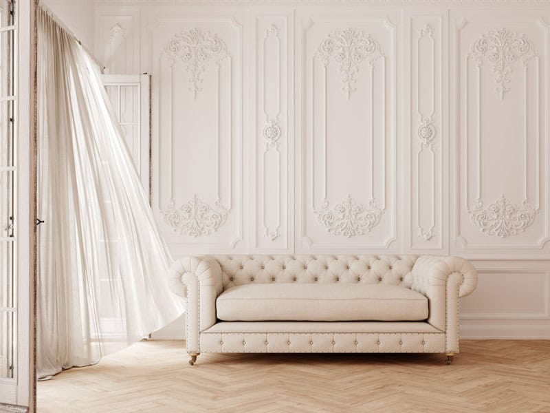 Elegant interior with a cream tufted sofa, ornate white wall panels, and a sheer curtain billowing near an open window, suggesting a serene ambiance.