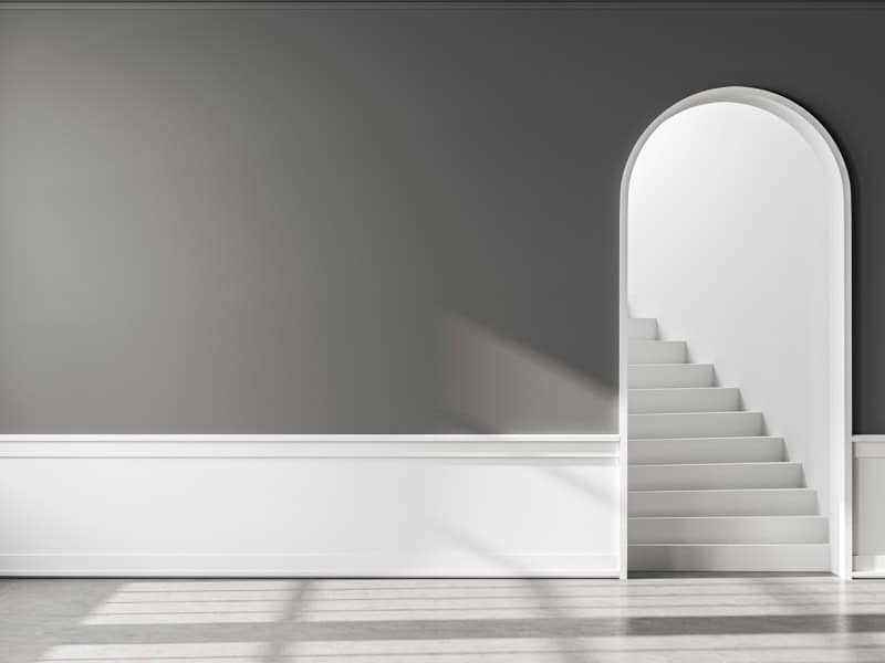 This image depicts a minimalist interior with an arched doorway leading to a white staircase, against a grey wall with sunlight casting sharp shadows.