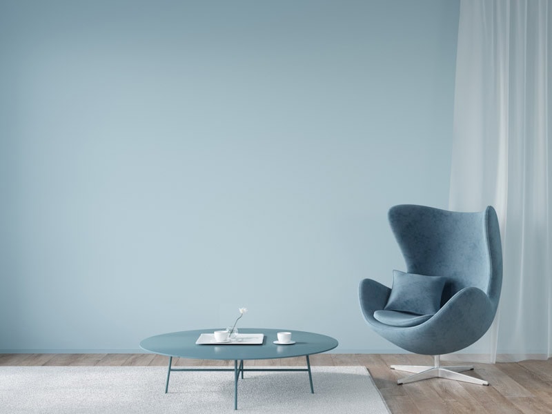 A minimalist room with a blue wall features a modern blue armchair, a round coffee table with a book and two cups, and sheer curtains.
