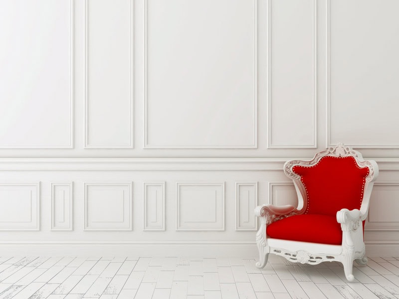 Elegant white room with classic paneling on walls, ornate white ceiling moldings, and a luxurious red velvet armchair with white ornate frame.