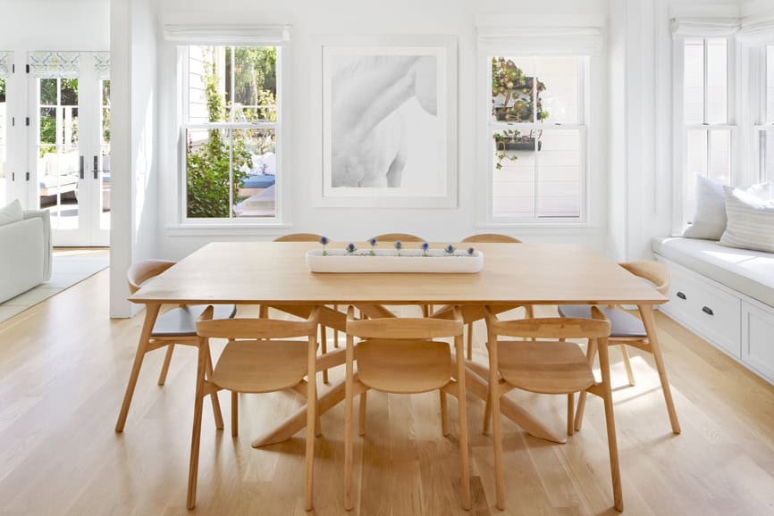 Bright, airy dining room with Scandinavian-style wooden table and chairs, white bench with cushions, light hardwood floor, artwork, and large windows.