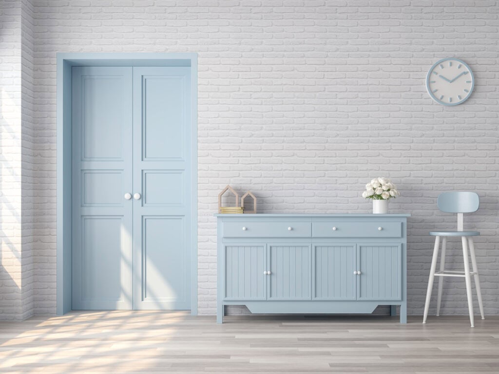A minimalist interior with a light blue door, matching sideboard, a clock, stool, and flowers against a white brick wall with natural light.