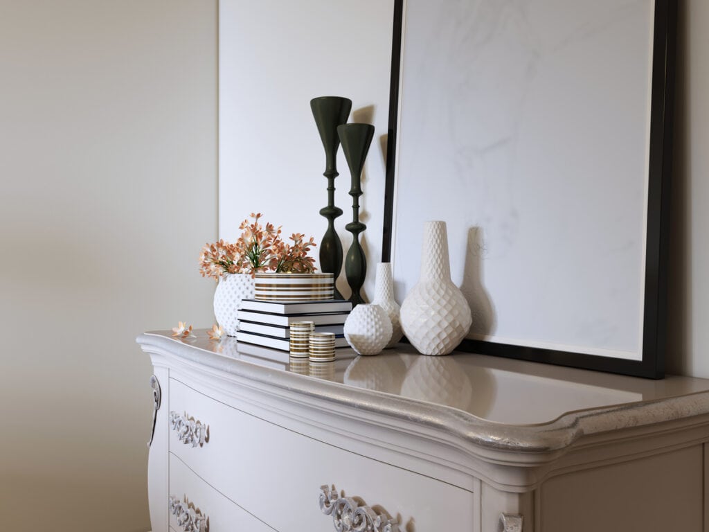 Elegant interior decor on a white vintage dresser featuring an assortment of decorative vases, books, and flowers next to a large framed mirror.
