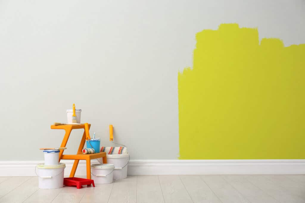 An unfinished bright yellow painting project on a wall with paint buckets, rollers, and a stool, suggesting a fresh coat of paint is an easy budget-friendly way to decorate your home.