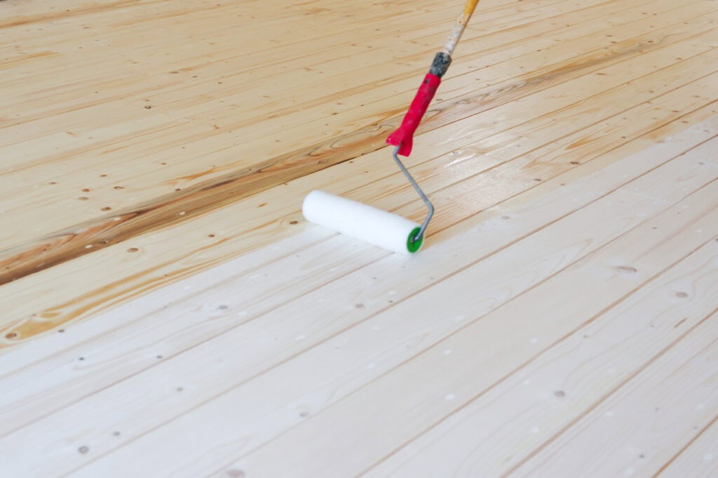 A paint roller with a red and black handle applies finish to a natural wooden floor. The image captures a home improvement project on a budget.