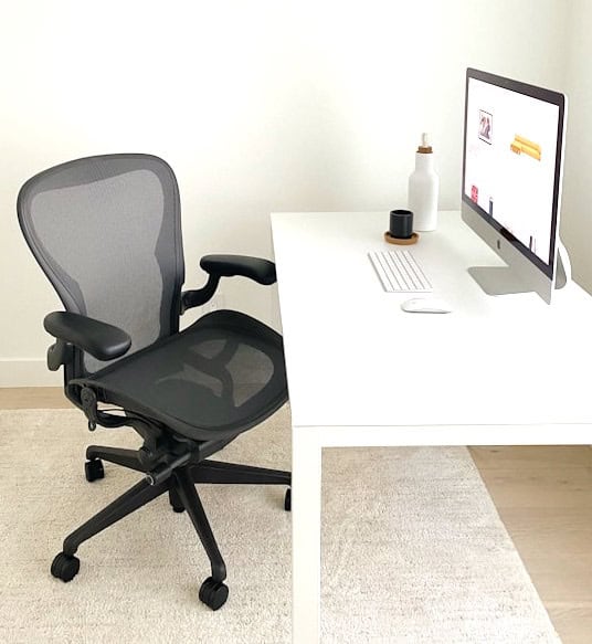 A modern, minimalist design home office with an ergonomic chair, white desk, laptop, mouse, monitor, and decorative bottle, on a white and beige rug.