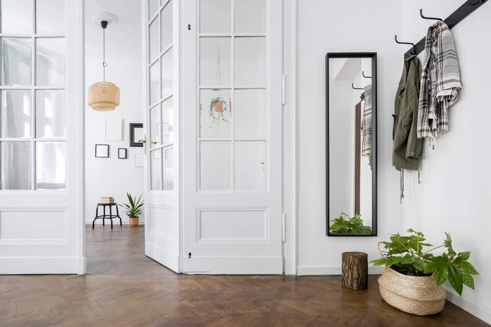 A bright, spacious entrance with herringbone wood flooring, white walls, potted plants, a hanging pendant light, and a coat rack next to a tall mirror.