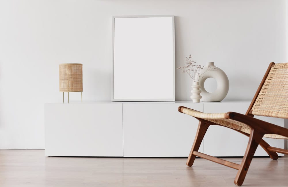 A minimalist room with a white wall, featuring a blank frame, a lamp, a decorative vase with dried plants, and a woven chair.