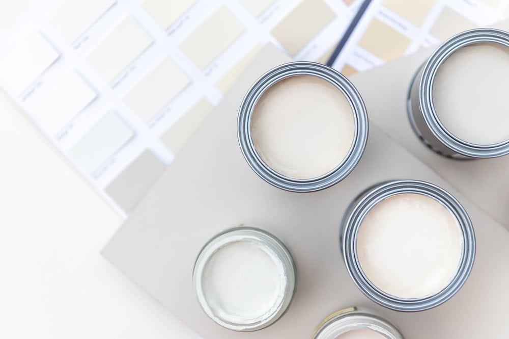 Top view of open paint cans with neutral colors, surrounded by paint swatches, on a white surface, depicting a modern minimalist house palette selection for interior design.
