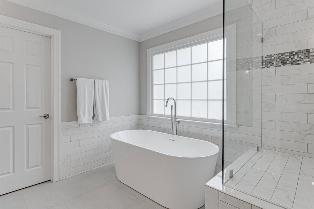 A modern minimalist bathroom with a freestanding tub, large frosted window, tiled shower area, white walls, and a towel hanging on the rack.