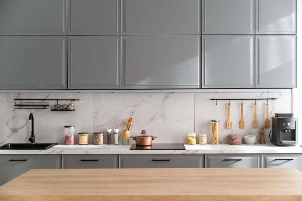 A modern kitchen with grey cabinets, marble backsplash, wooden utensils, and jars on the countertop. A pot, sink, and appliances are visible.