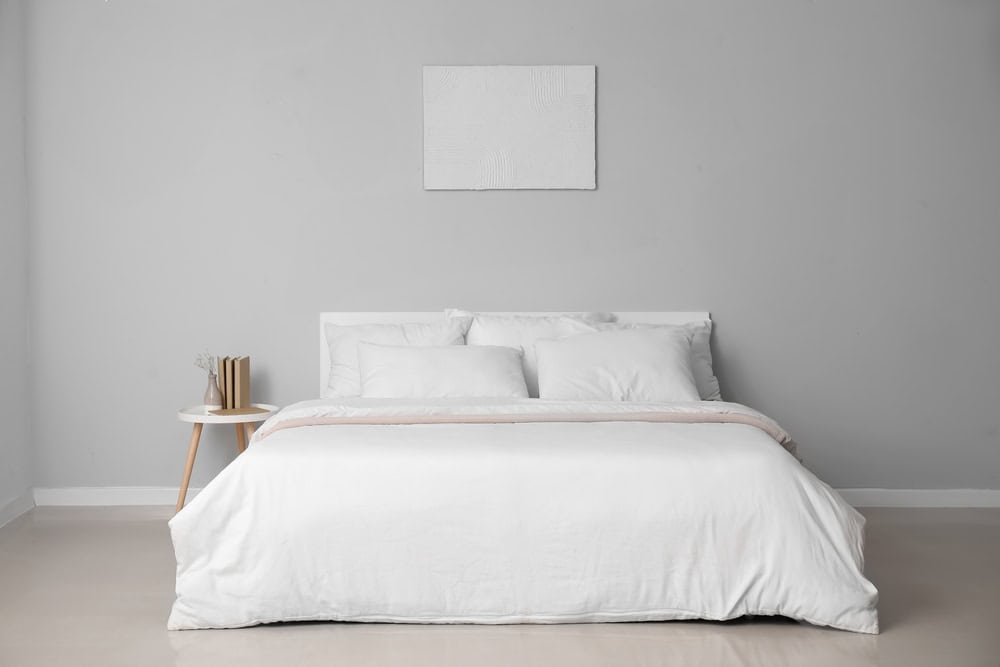 This is a minimalist bedroom with a neatly made bed, white bedding, a small round side table with books, and a textured painting above the bed.
