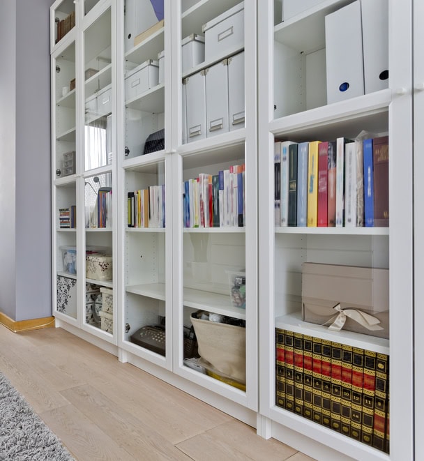 The image shows a modern, organized white bookshelf with books, storage boxes, and folders in a room with a gray wall and wooden floor. Installed in a way that makes the furniture look built-in - an excellent example of home decor on a budget.