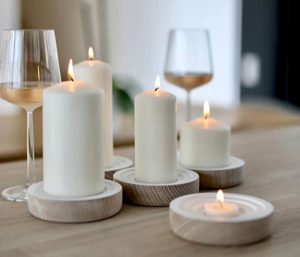 The image displays a serene modern minimalist design setting with several lit candles of various sizes on wooden holders, accompanied by two glasses of white wine.