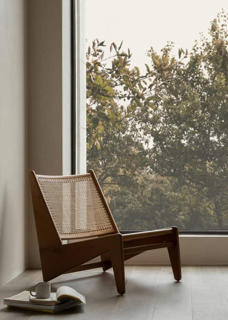 A wooden chair with woven seat is placed by a window, facing a view of trees in a modern minimalist house design. A cup and open book rest on the floor beside it.