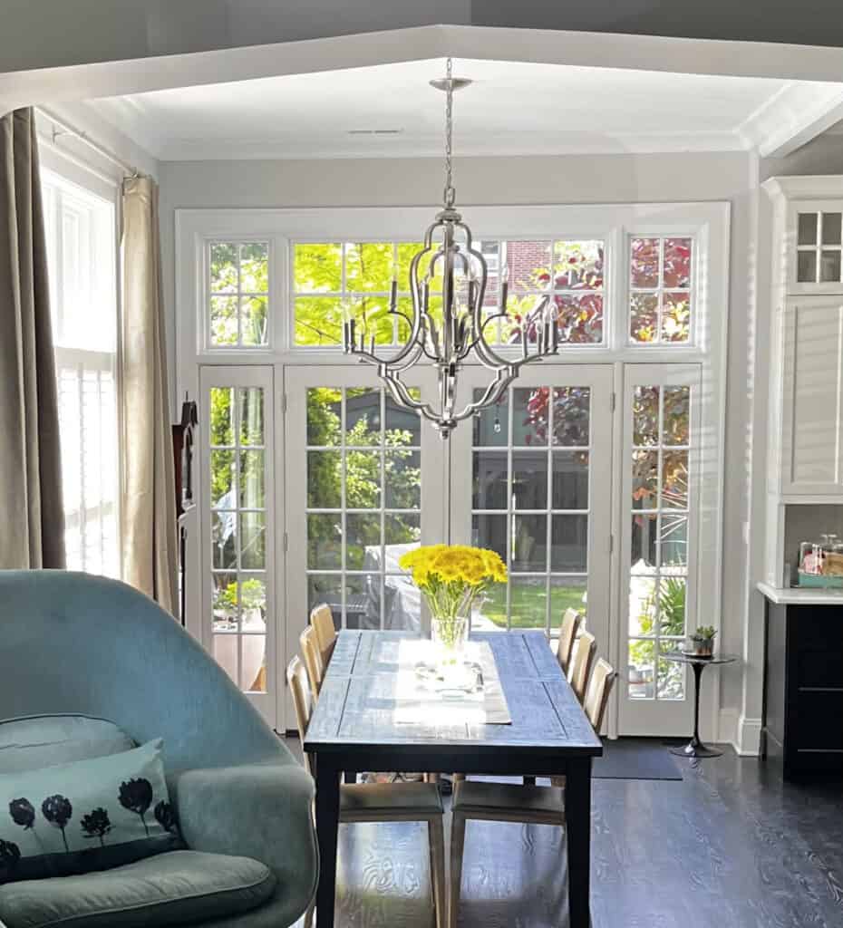A bright dining area with a table, chairs, yellow flowers, large windows, and a chandelier, opening to a lush garden.